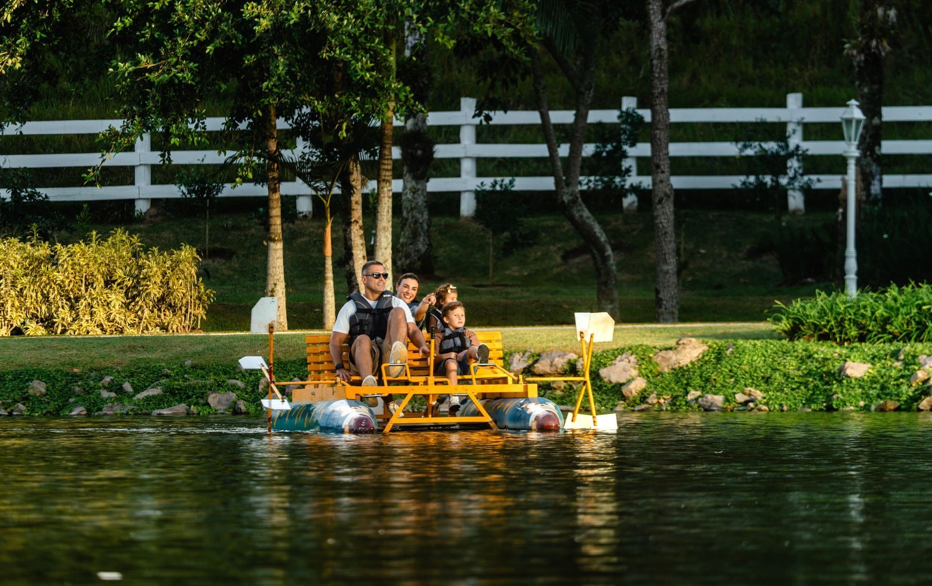 Duas pessoas remando em uma canoa vermelha em um lago com uma cerca branca e árvores verdes ao fundo.