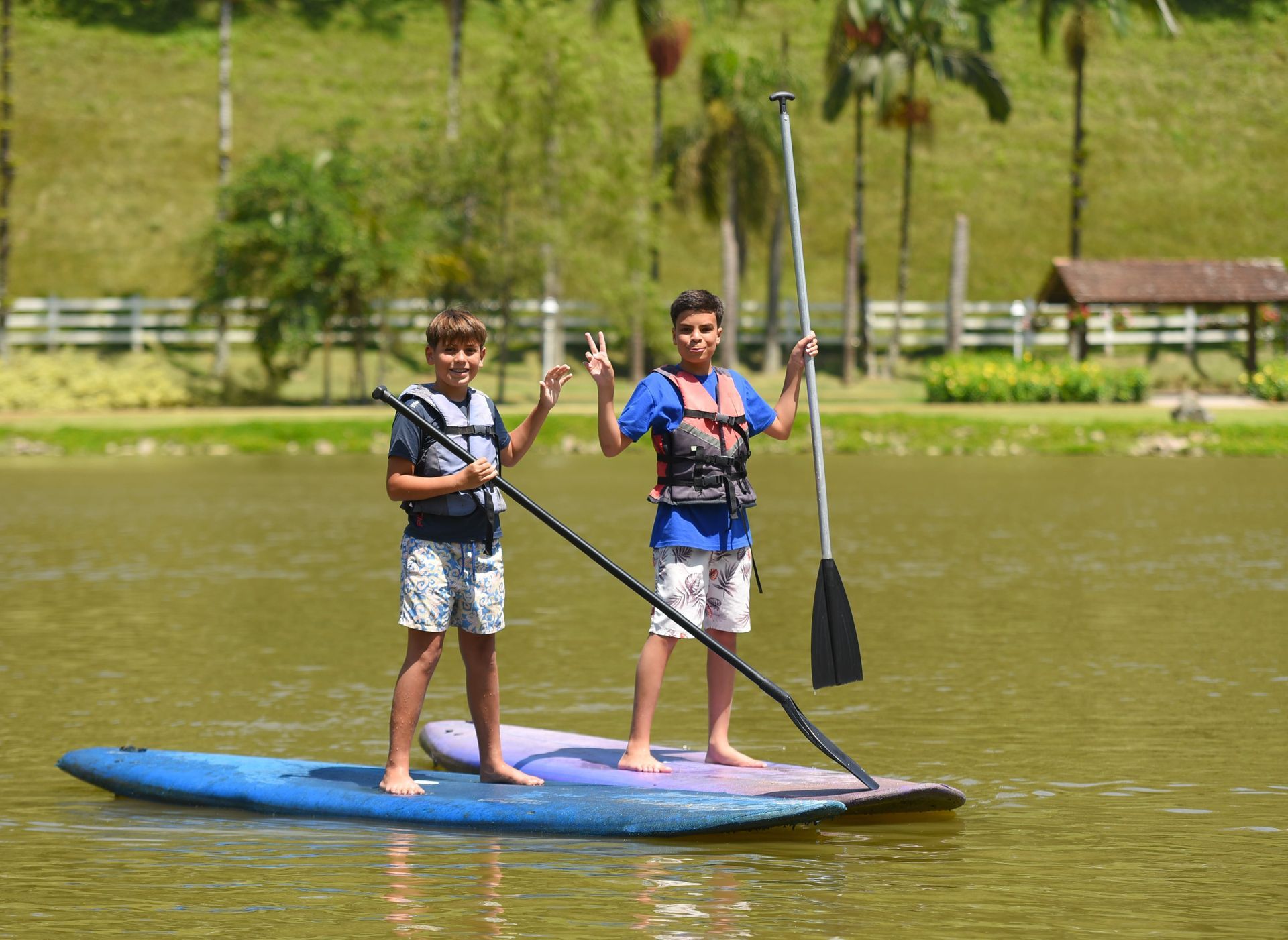 Pessoa praticando stand-up paddle em um lago, segurando um remo e com um braço levantado em um parque arborizado.