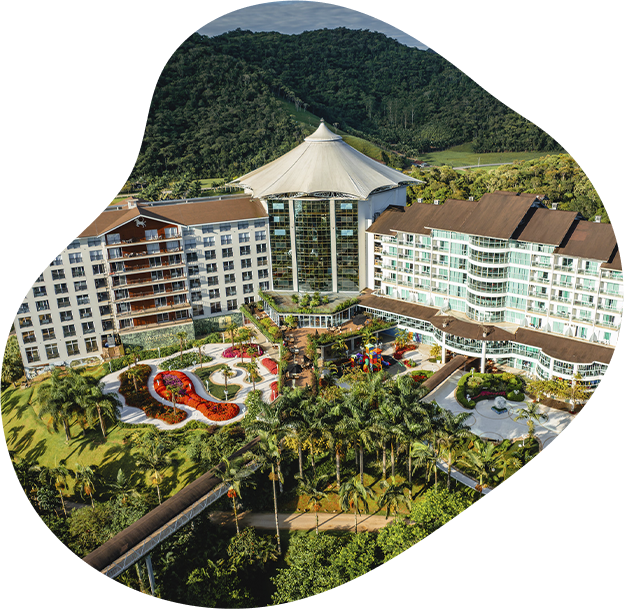 Vista aérea de um resort tropical com edifícios brancos curvos, palmeiras, piscinas e montanhas verdes ao fundo.