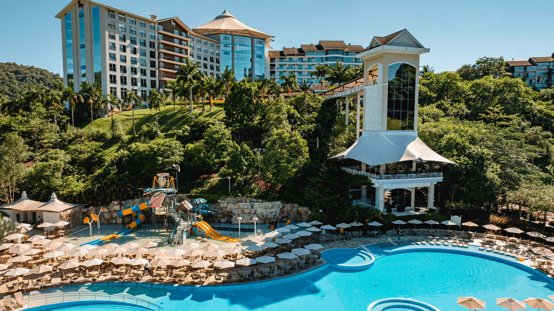 Piscina do resort com água turquesa, paisagismo tropical e altos edifícios de hotel à beira-mar.