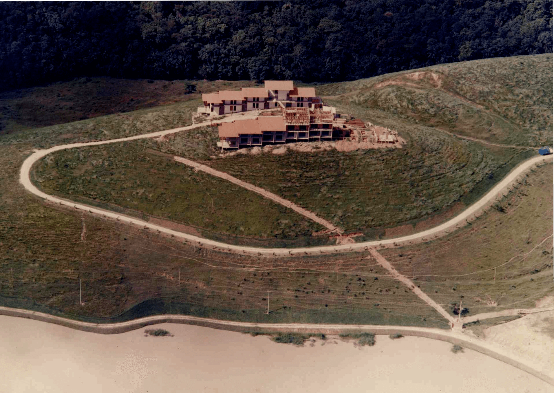 Vista aérea de uma casa em uma colina gramada com caminhos de terra sinuosos perto de uma costa arenosa.