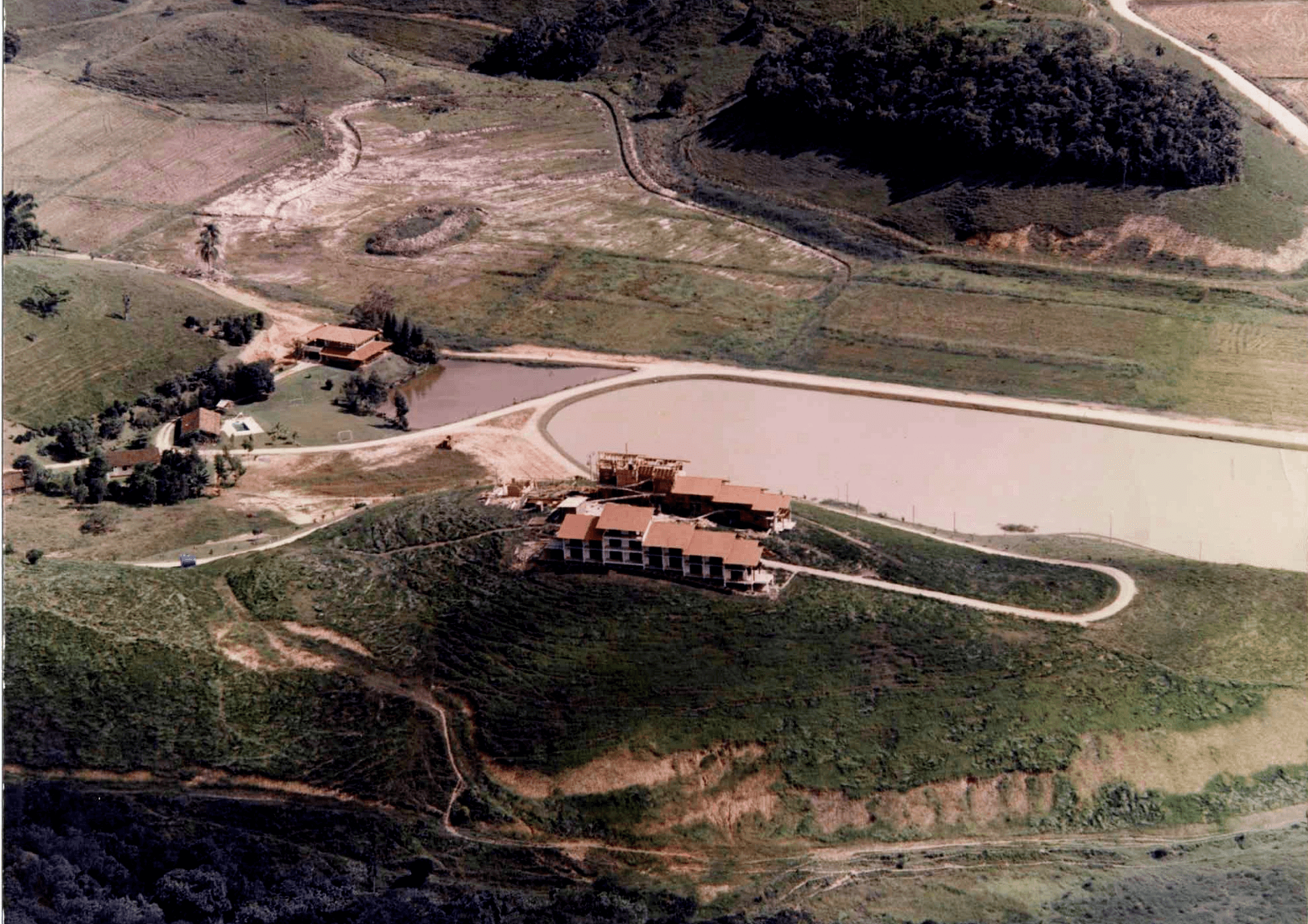 Vista aérea de uma casa ao lado de um lago, cercada por colinas gramadas e estradas de terra sinuosas.