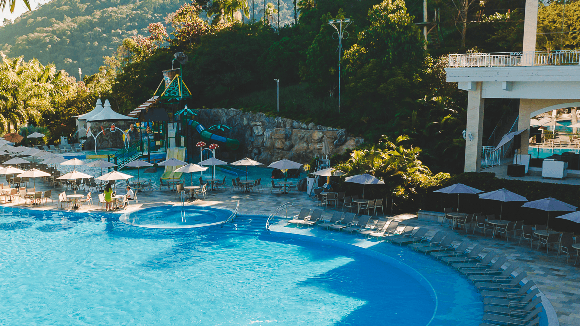 Piscina de resort com água azul, espreguiçadeiras, guarda-sóis e vegetação tropical ao lado de um edifício.