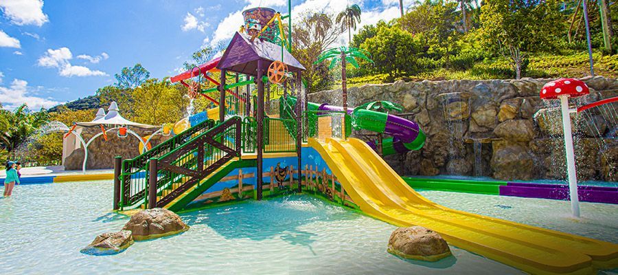 Parque aquático colorido com escorregadores amarelos, estruturas de brincar e piscina rasa em um parque ensolarado ao ar livre.