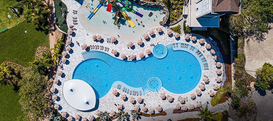 Vista aérea da piscina de um resort com água azul, espreguiçadeiras brancas, guarda-sóis e palmeiras ao redor.