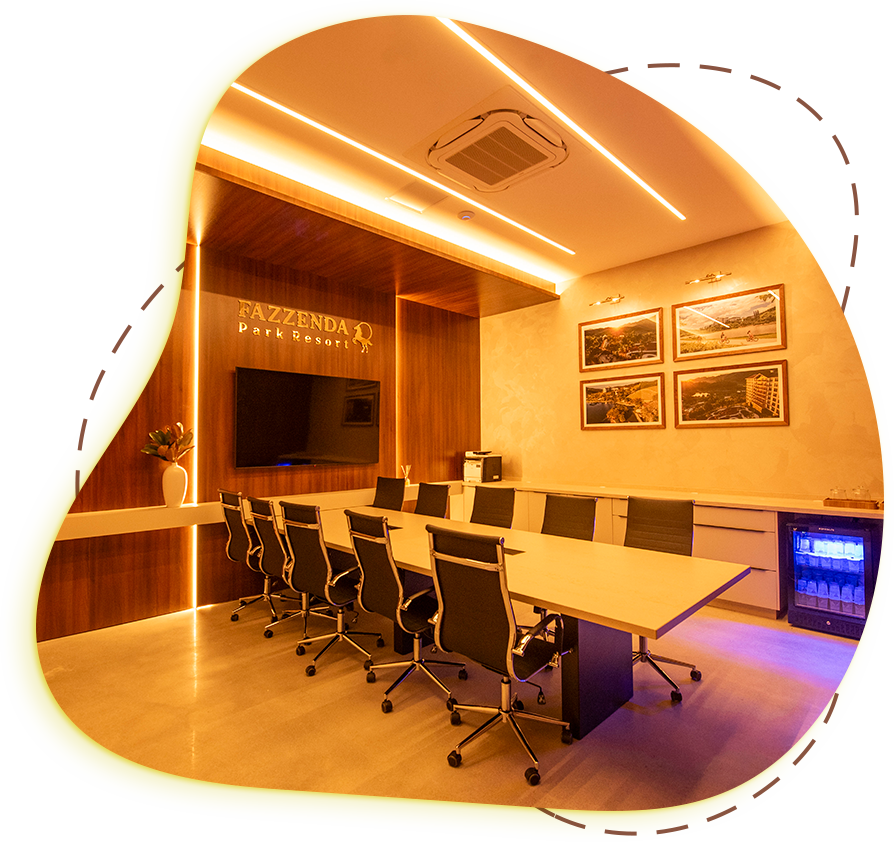 Sala de conferências moderna com iluminação aconchegante, mesa oval, cadeiras pretas, TV fixada na parede e fotos emolduradas.