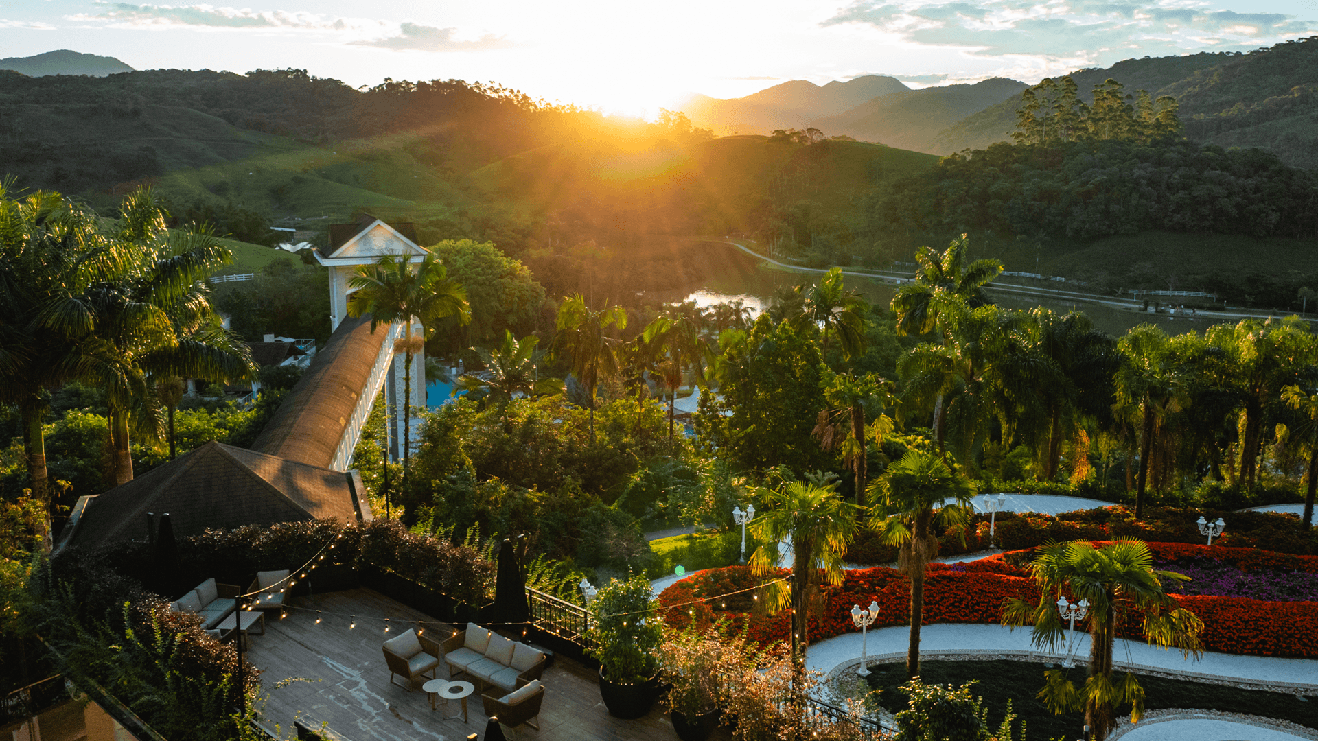 Pôr do sol sobre um resort tropical com palmeiras, jardins, piscinas e uma ponte que leva às colinas.