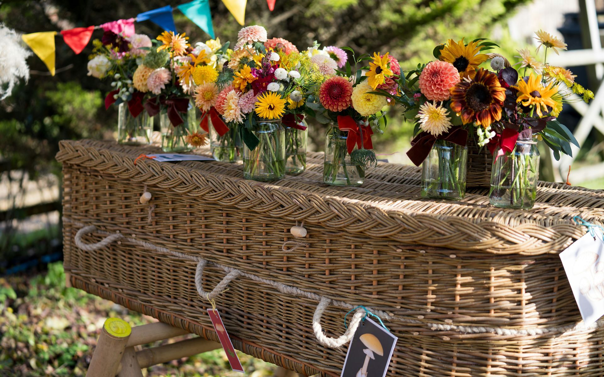 Wicker coffin with floral arrangements in jars. Colorful garland in background.