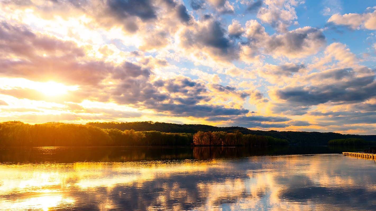 Sunset over a calm lake, reflecting golden light and cloudy sky; treeline on the horizon.