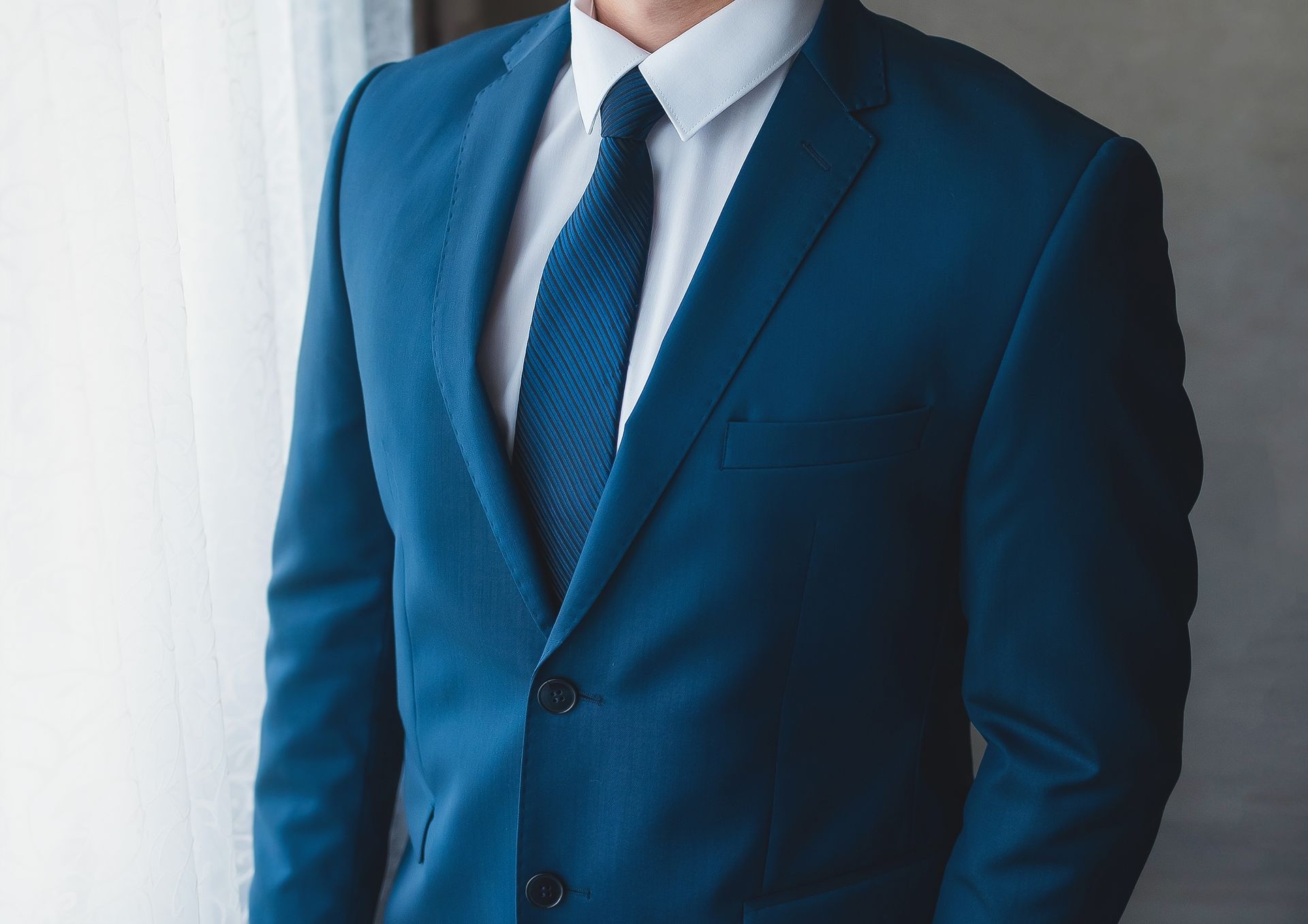 Man in a blue suit, white shirt, and blue tie, standing indoors near a window.