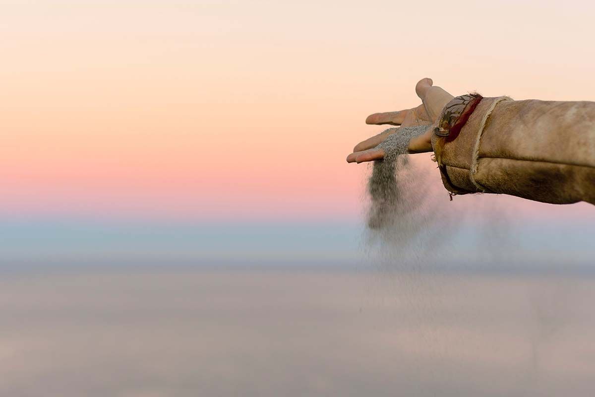 Hand releasing sand against a pastel sky over a body of water.