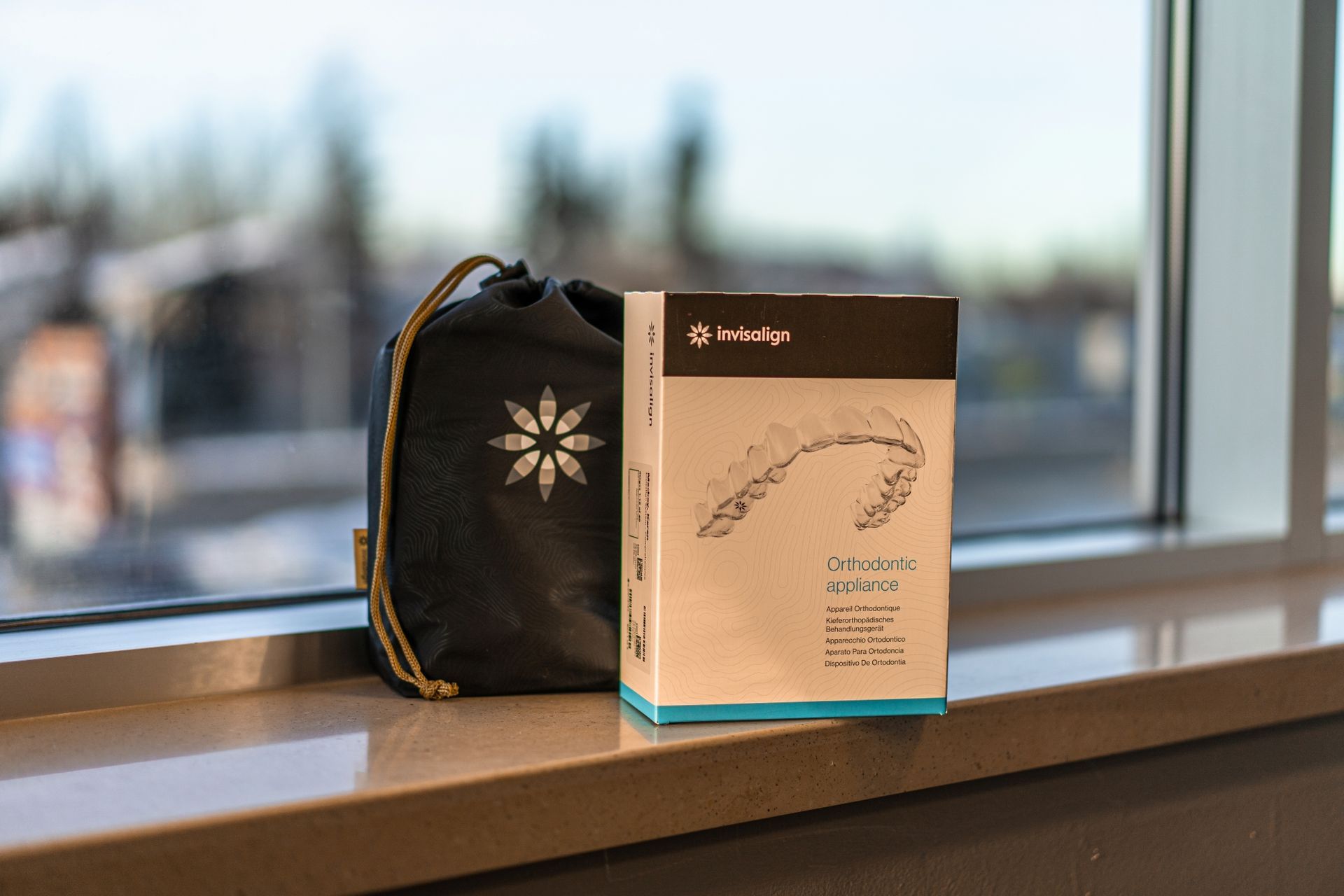 A black drawstring bag with a white flower logo and a white box for a mandibular appliance sit on a windowsill.