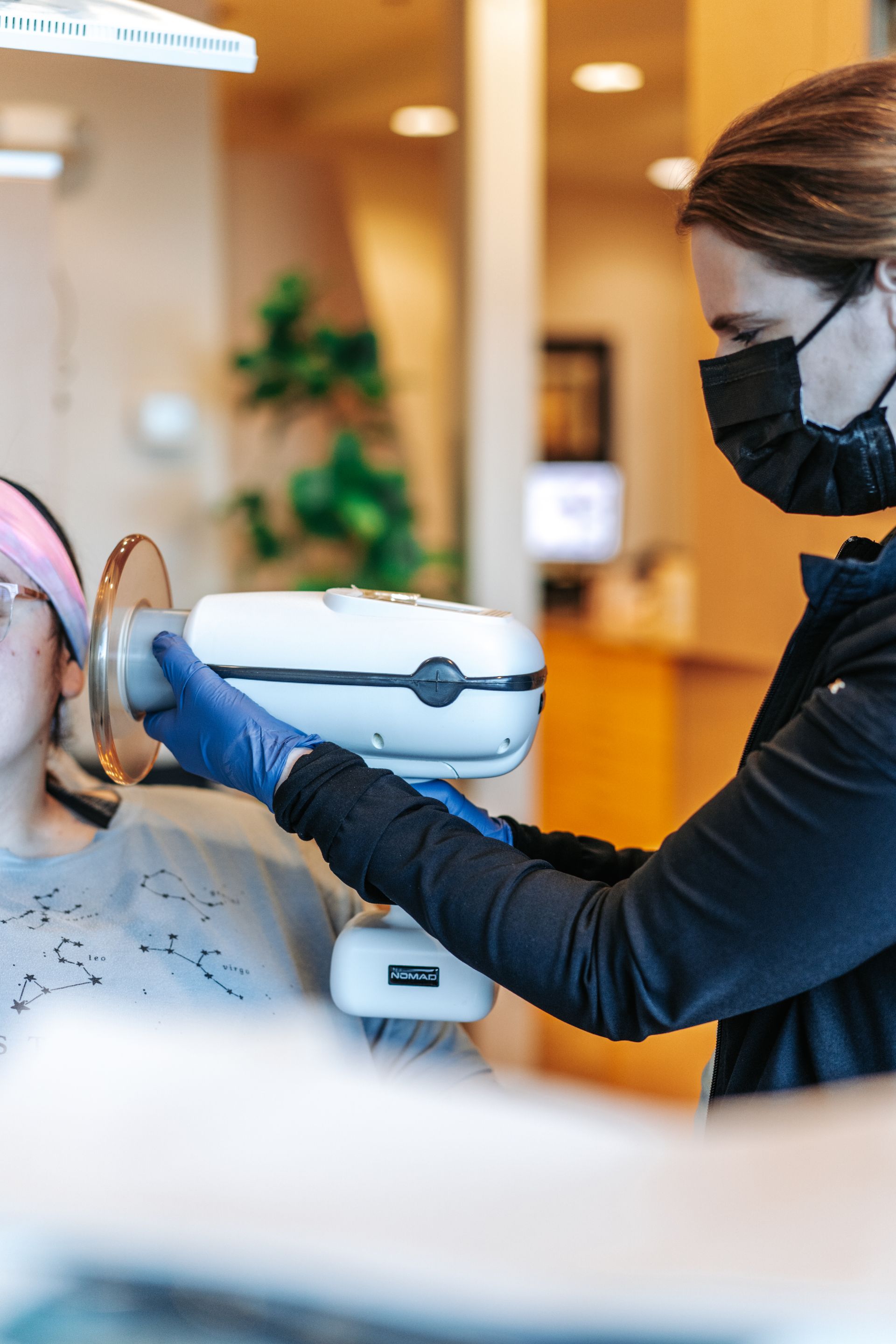 A dental professional in a mask and blue gloves uses a handheld X-ray device on a patient in a clinical office setting.