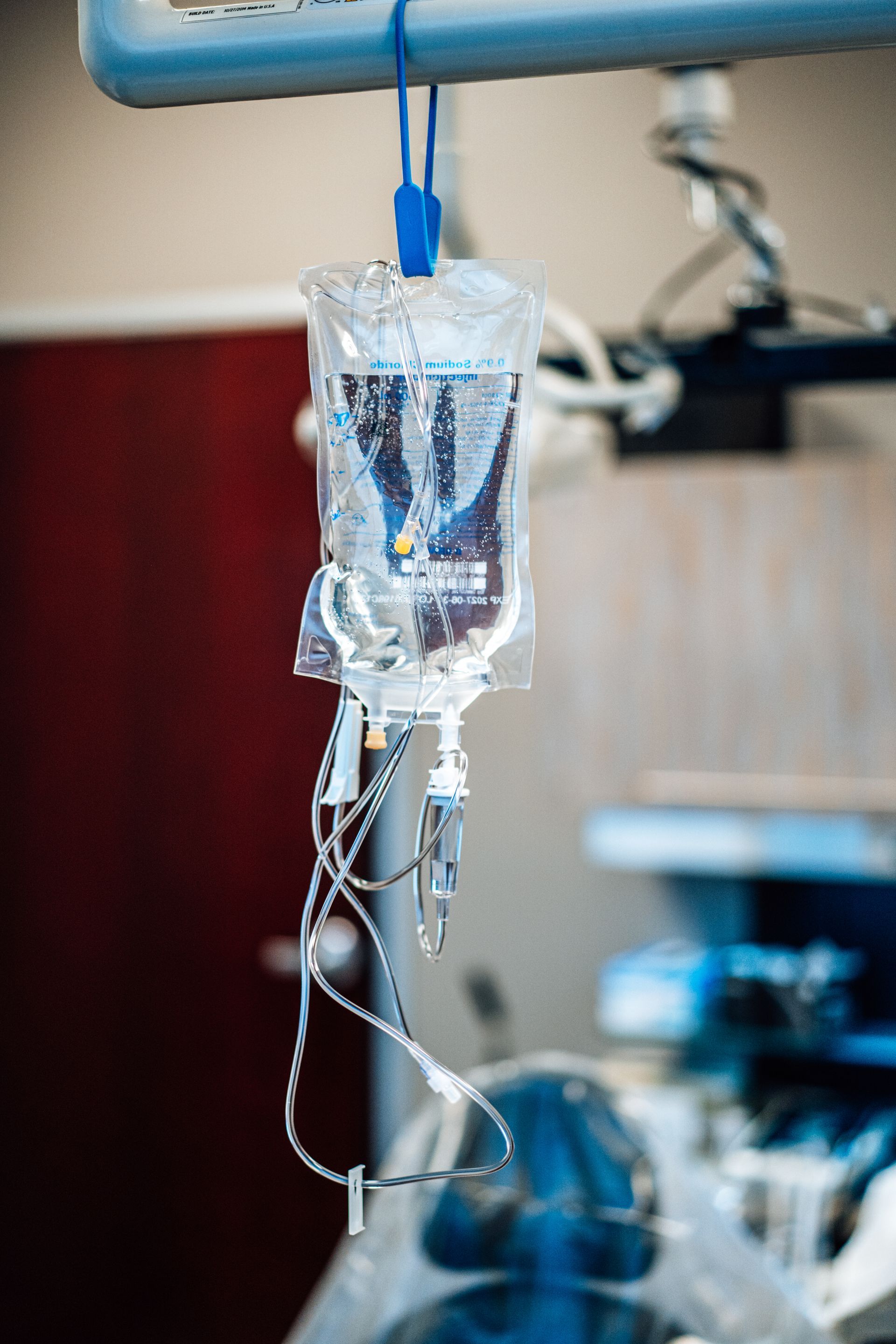 A clear medical IV fluid bag with tubing hanging from a stand in a clinical, out-of-focus background.