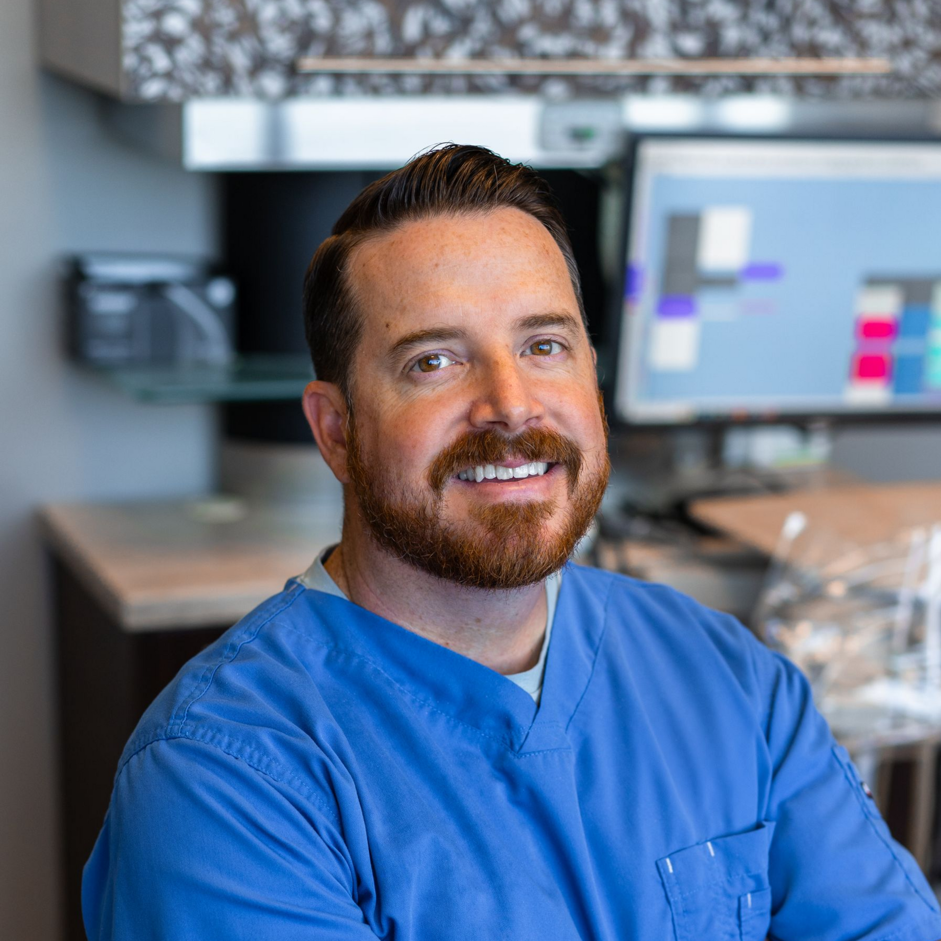 A professional in blue scrubs smiling in an office setting with a computer monitor in the background.