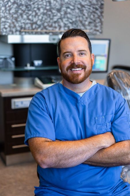 Man in blue scrubs with arms crossed, smiling in a dental office setting.