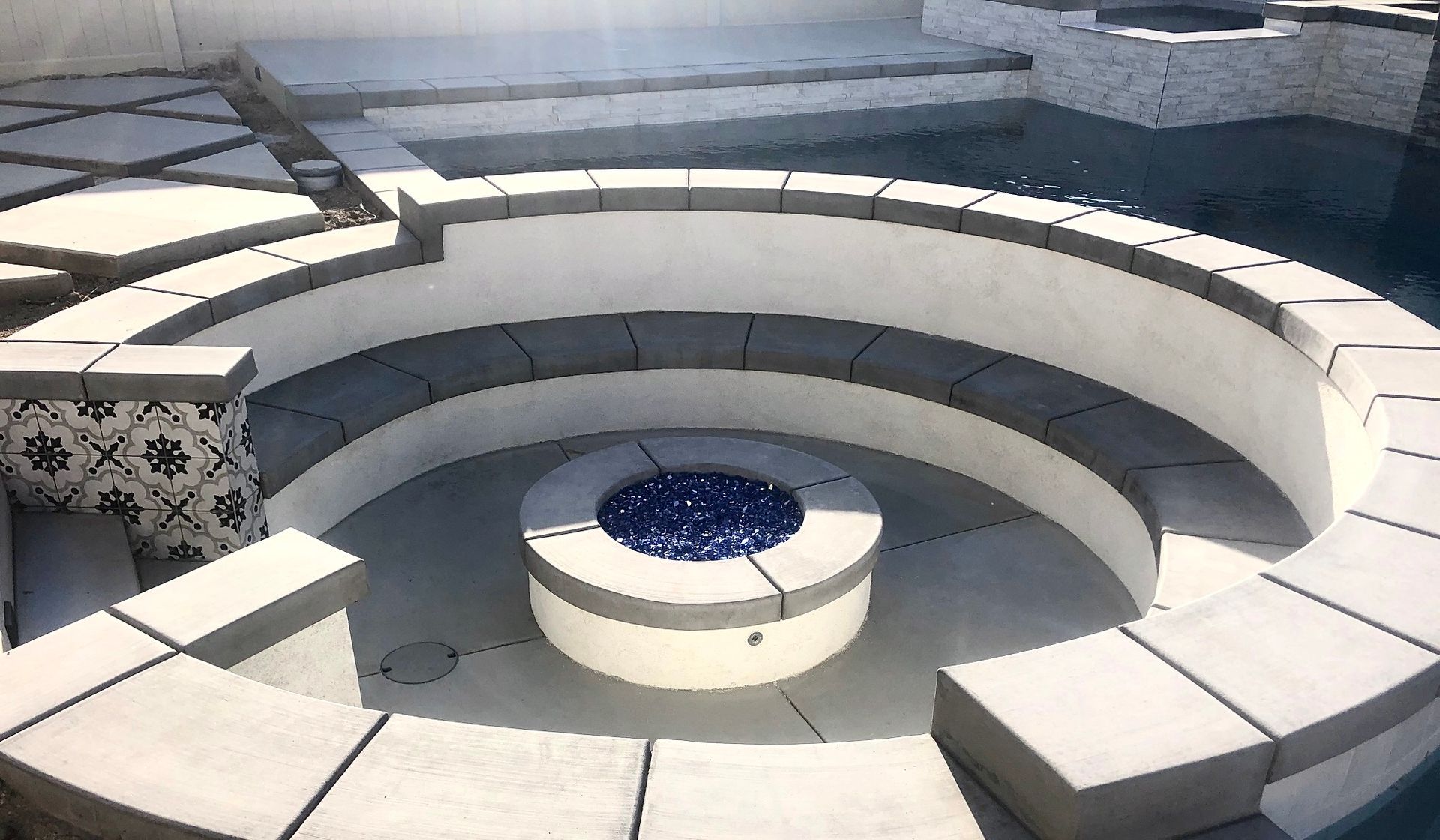 A sunken circular stone fire pit with a blue glass center, surrounded by curved white bench seating and tiled accents.