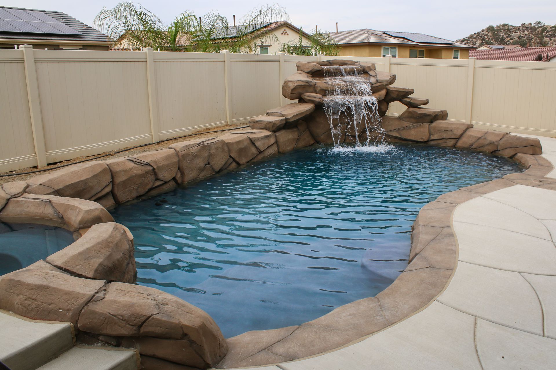 A backyard swimming pool with a rock-style waterfall feature surrounded by a tan vinyl fence and a concrete patio.