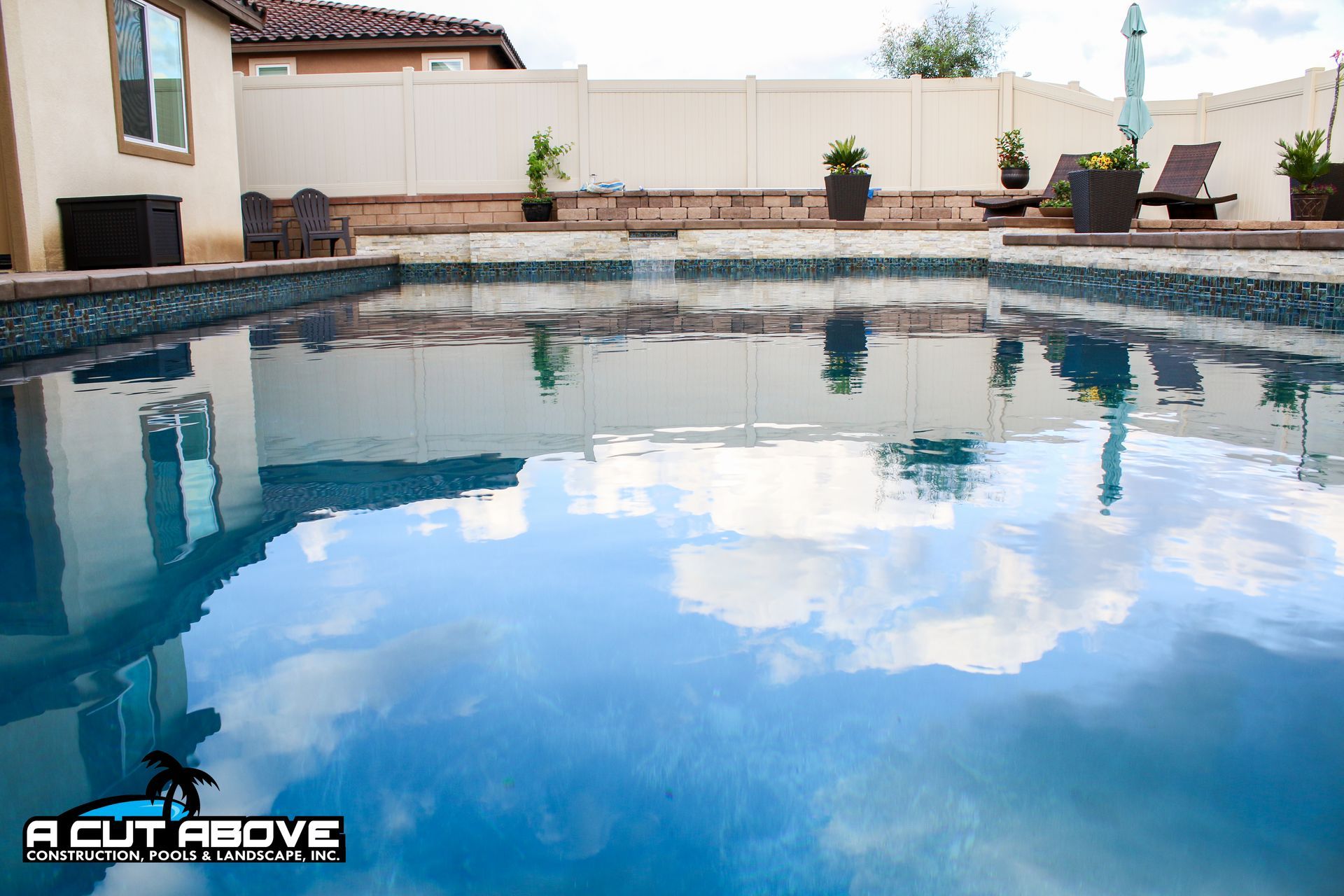 A clear swimming pool with reflections of the sky and house, set in a backyard with a fence and potted plants.