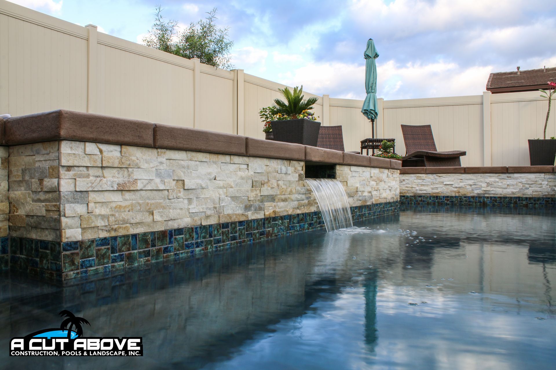 A swimming pool features a stone raised wall with a water feature, blue tile accents, lounge chairs, and a white fence.