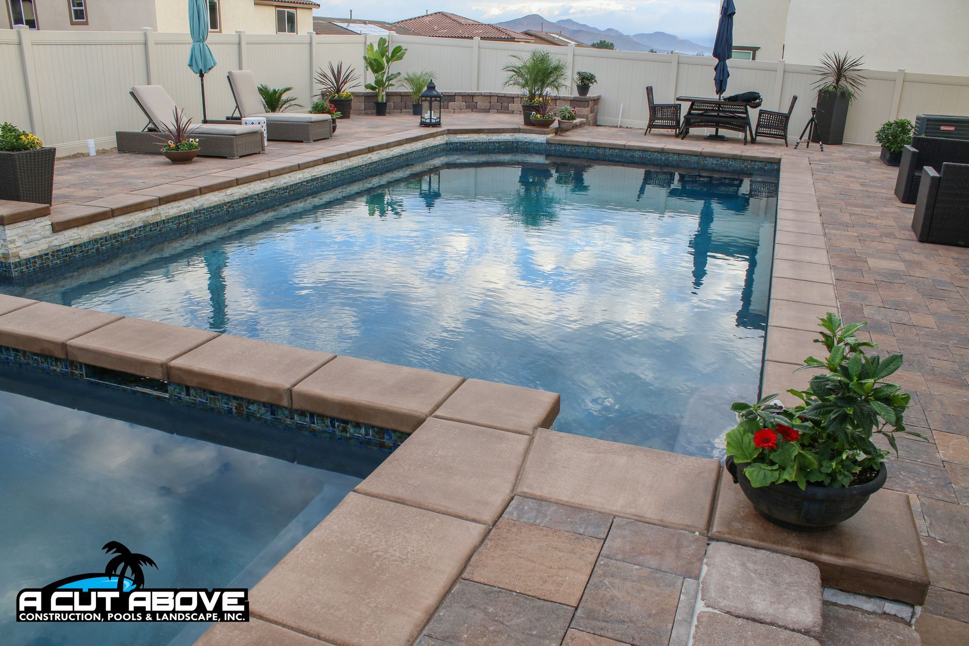 An outdoor pool area with tan paver decking, lounge chairs, a dining set, and potted plants against a white privacy fence.