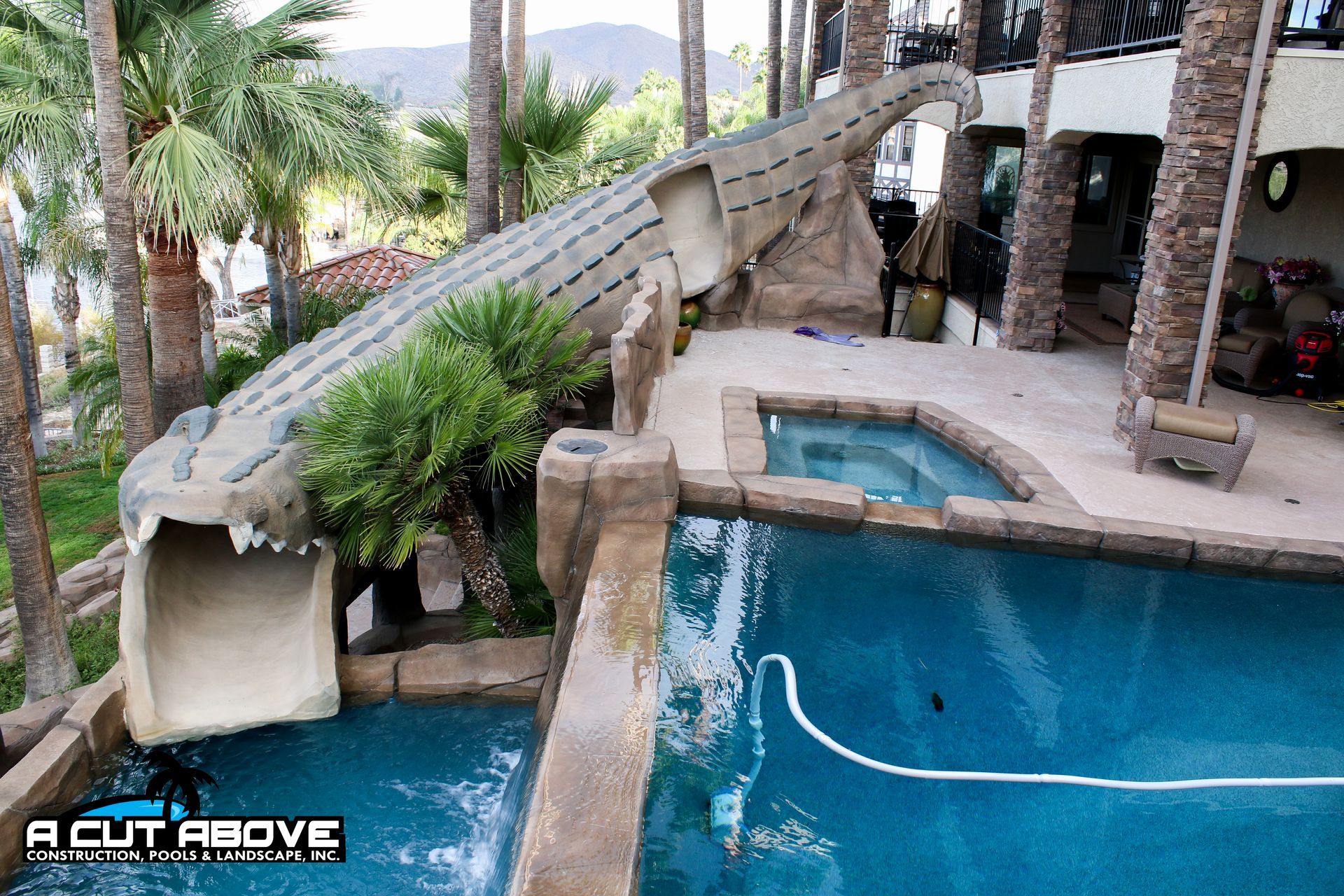 A stone alligator-shaped water slide leads into a backyard swimming pool next to a patio and a small hot tub.
