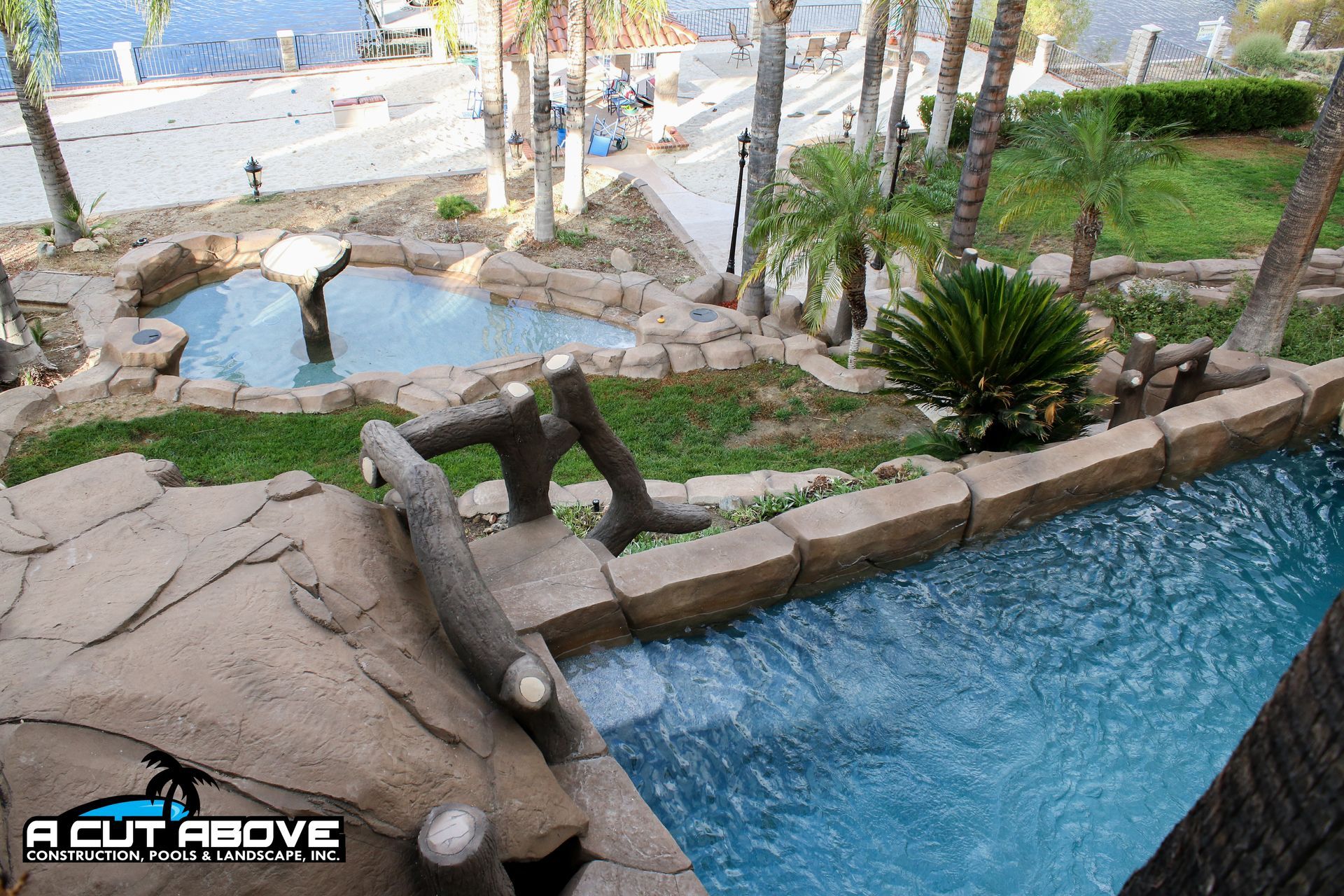 A high-angle view of a backyard featuring a swimming pool, a stone-bordered fountain, palm trees, and landscaped grass.