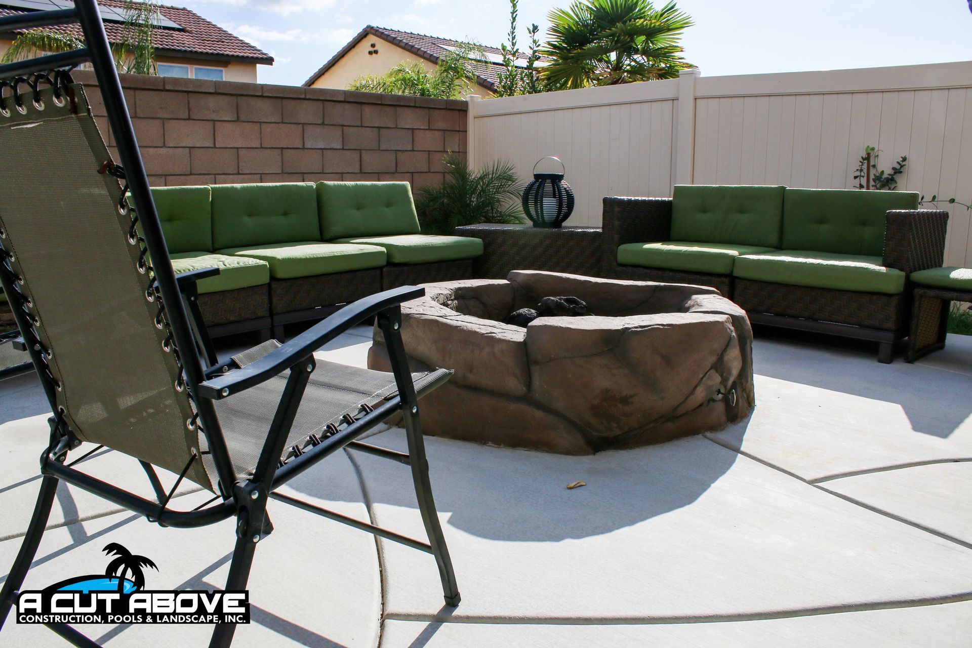A stone-look fire pit centered on a patio with two L-shaped wicker sofas with green cushions and a lawn chair.