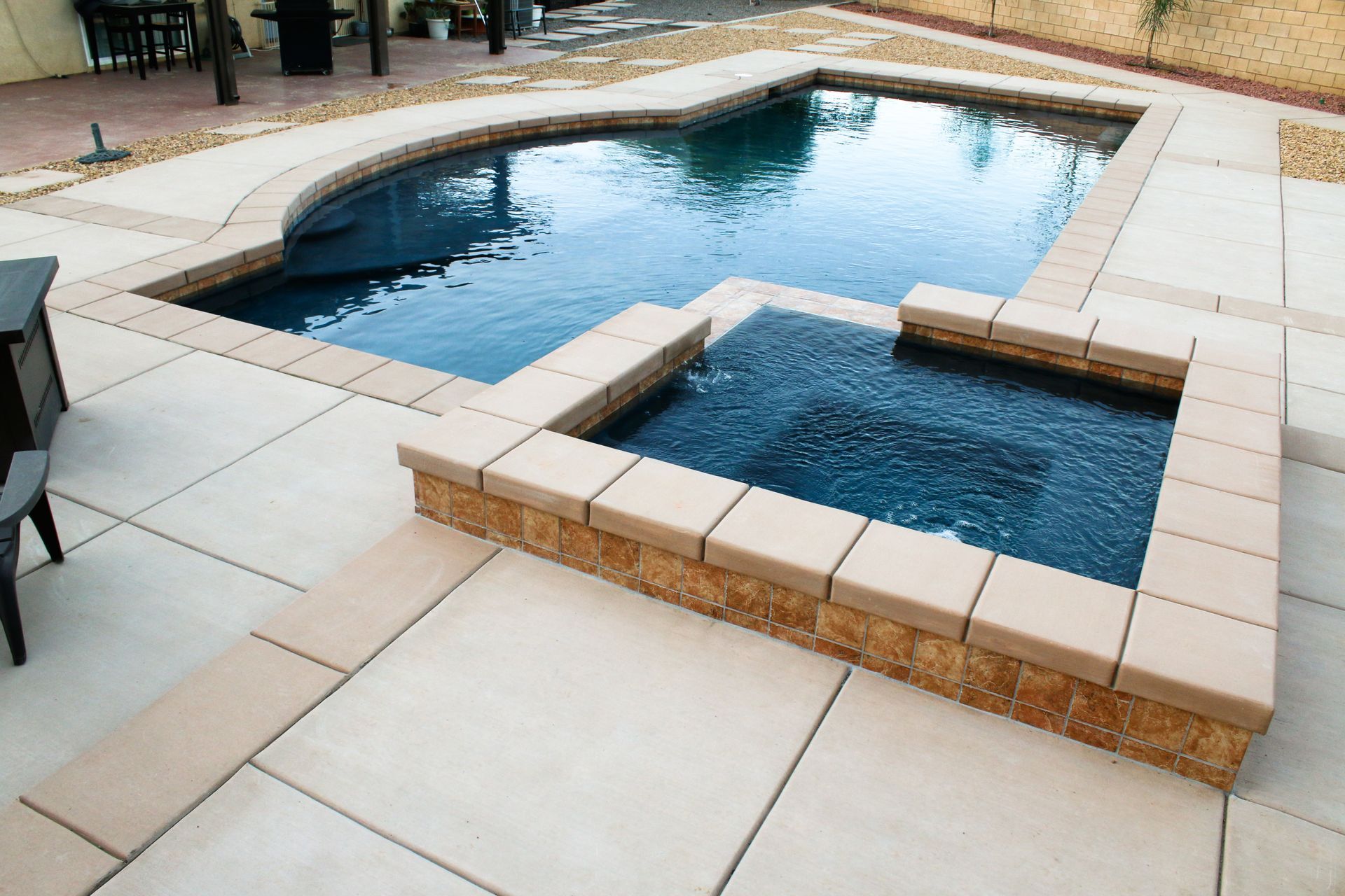 A backyard swimming pool with an attached square, raised hot tub featuring stone coping and light-colored concrete pavers.
