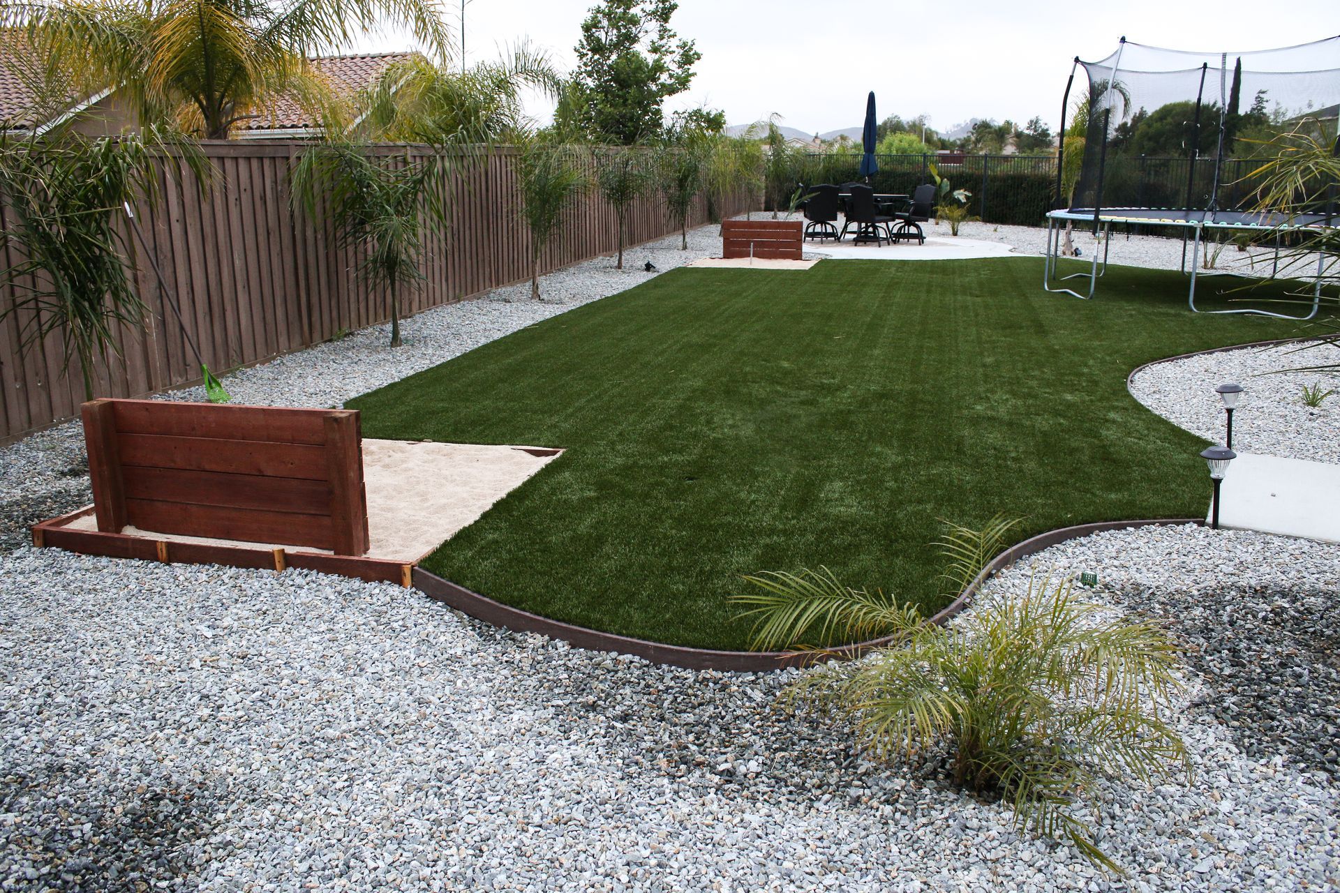 A landscaped backyard with a green artificial turf lawn, bordered by grey gravel, wooden raised beds, and a trampoline.