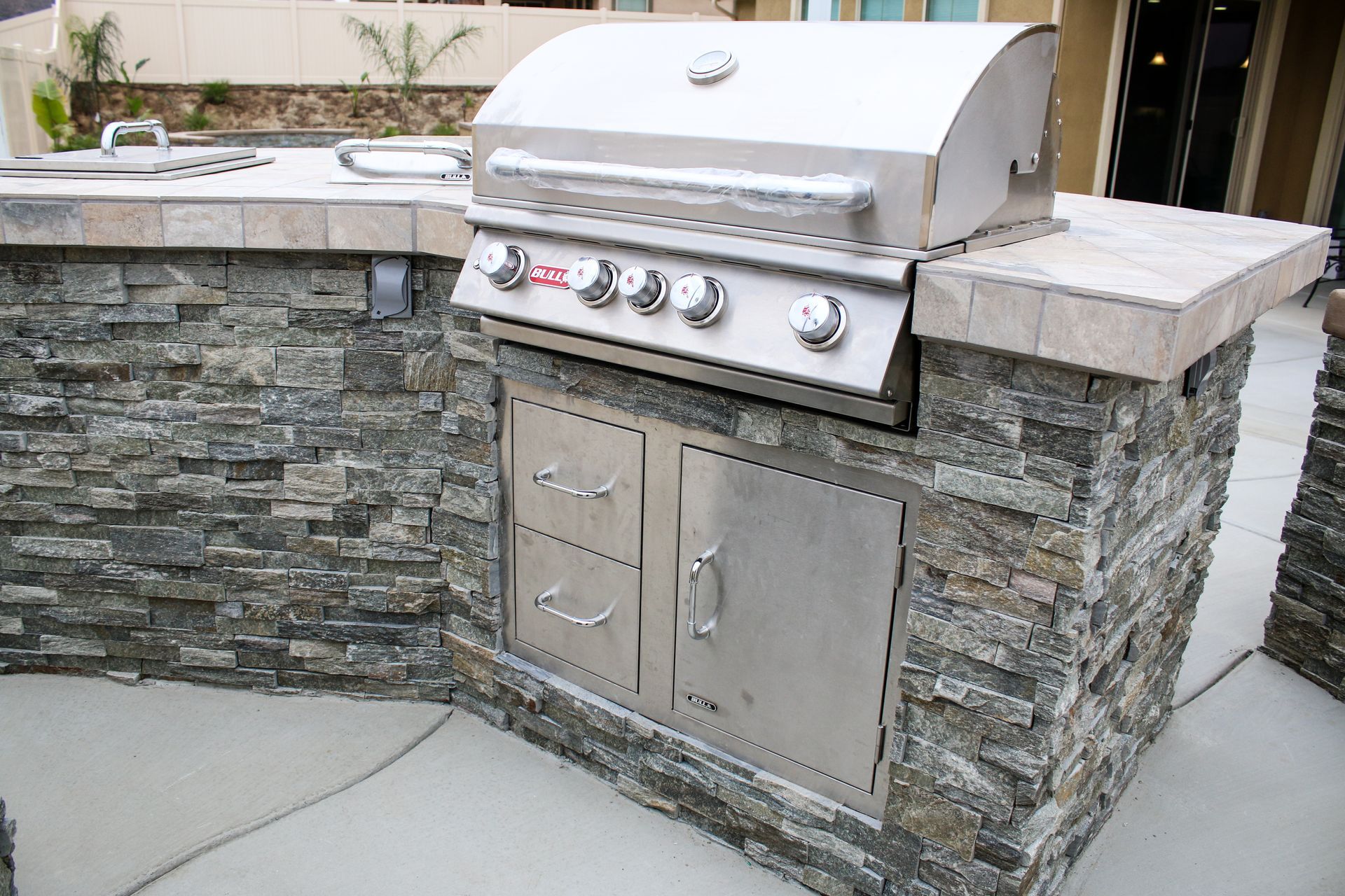 Stainless steel gas grill built into an outdoor stone counter with drawers and storage cabinet on a concrete patio.