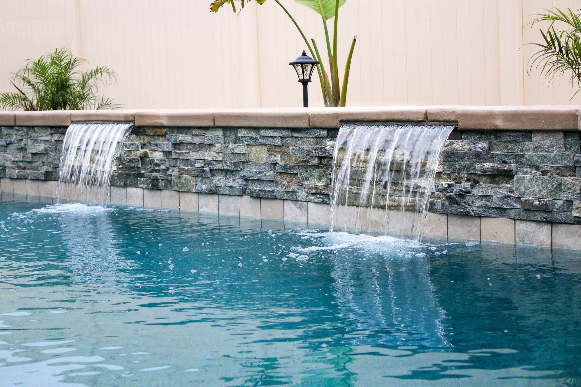 Two water features cascade from a stone wall into a blue swimming pool in a backyard setting.