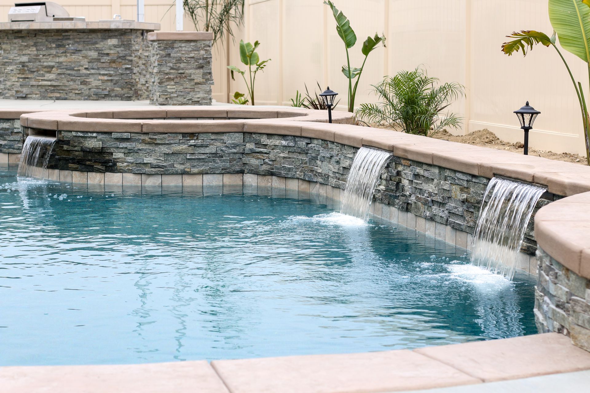 A pool with stacked stone walls and two waterfalls, featuring a raised hot tub and landscape lighting in a backyard.
