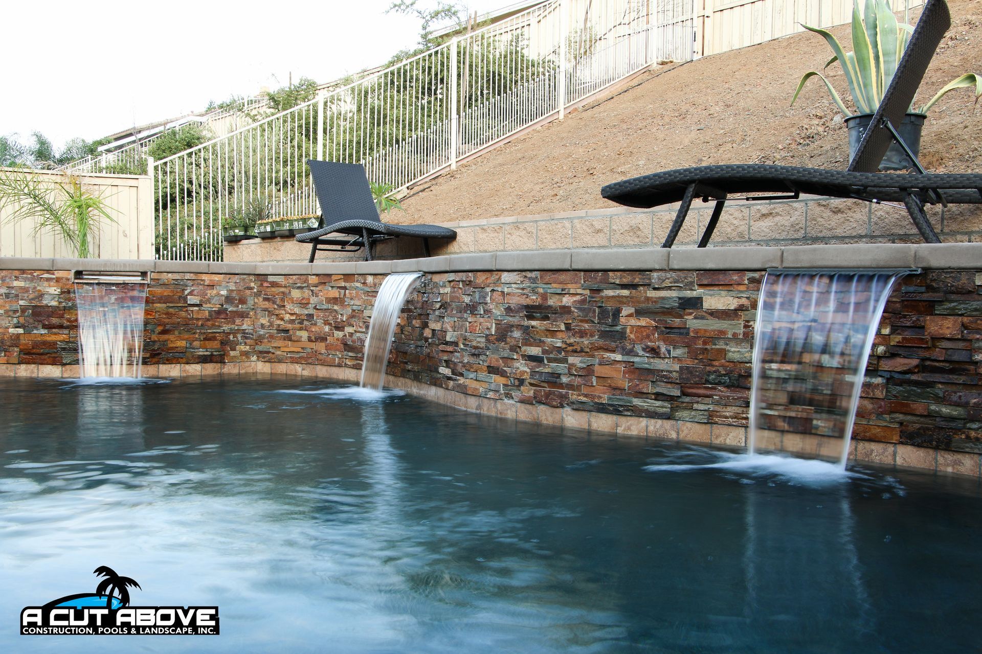 Three stone-walled waterfalls spill into a dark blue swimming pool, with lounge chairs on the patio above.