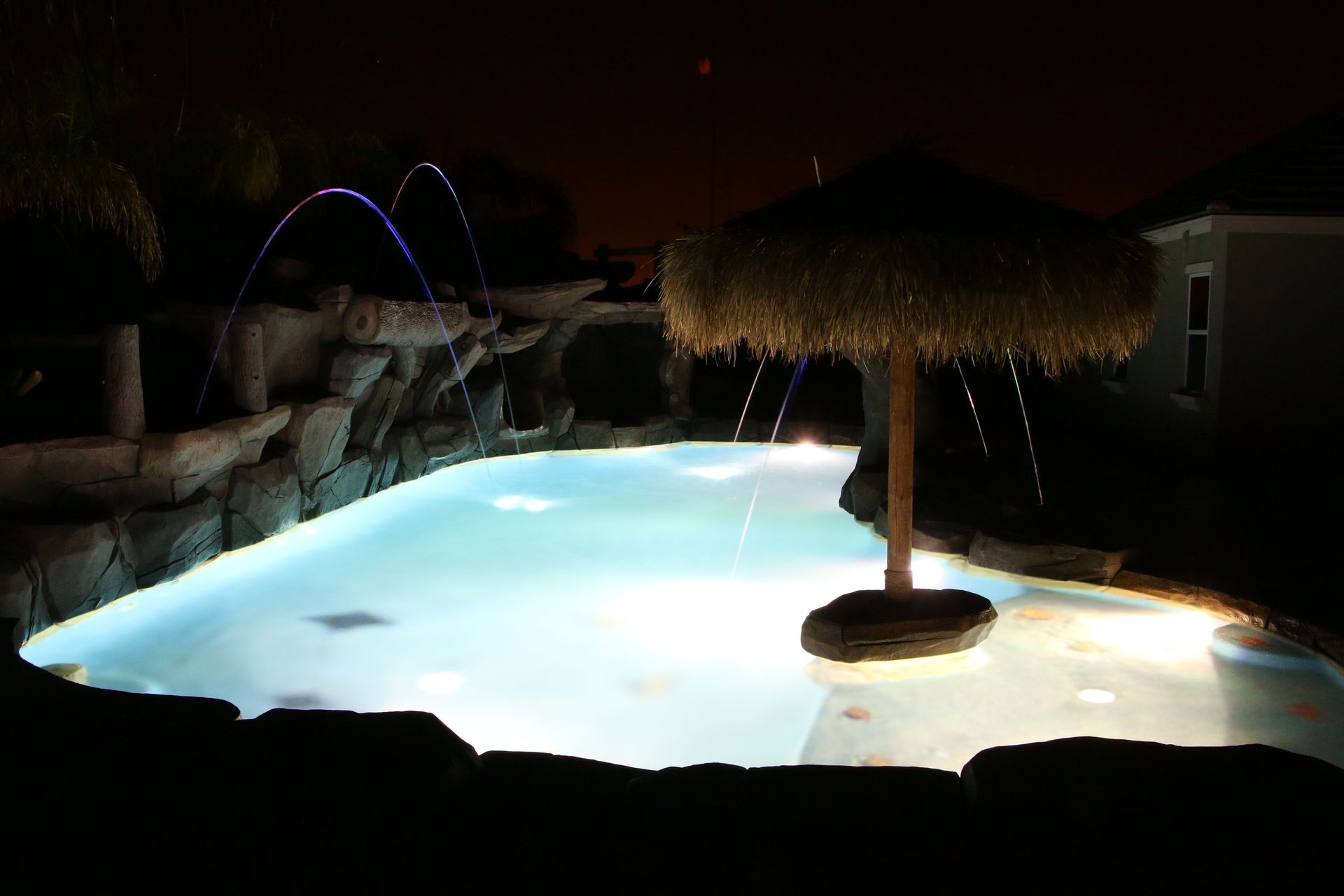 A swimming pool at night featuring glowing blue lights, a tiki umbrella in the shallow end, and illuminated water jets.