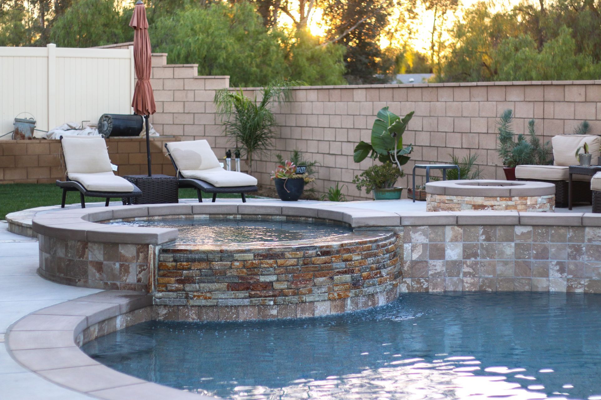 A backyard pool area with a stone hot tub, waterfall edge, lounge chairs, and a fire pit against a masonry wall at sunset.