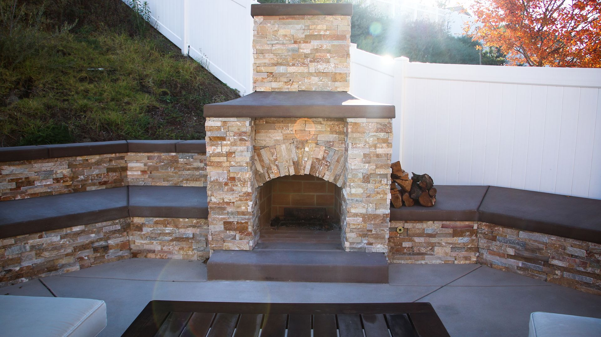 A stone outdoor fireplace with curved built-in seating on a patio against a white fence.