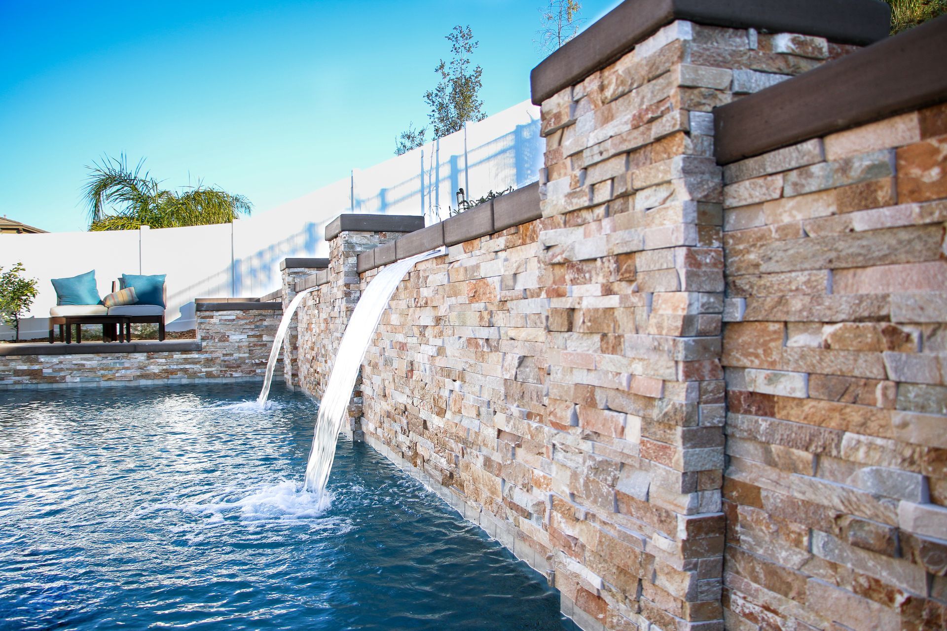 A stone retaining wall with two water features flowing into a backyard pool, with lounge seating in the background.