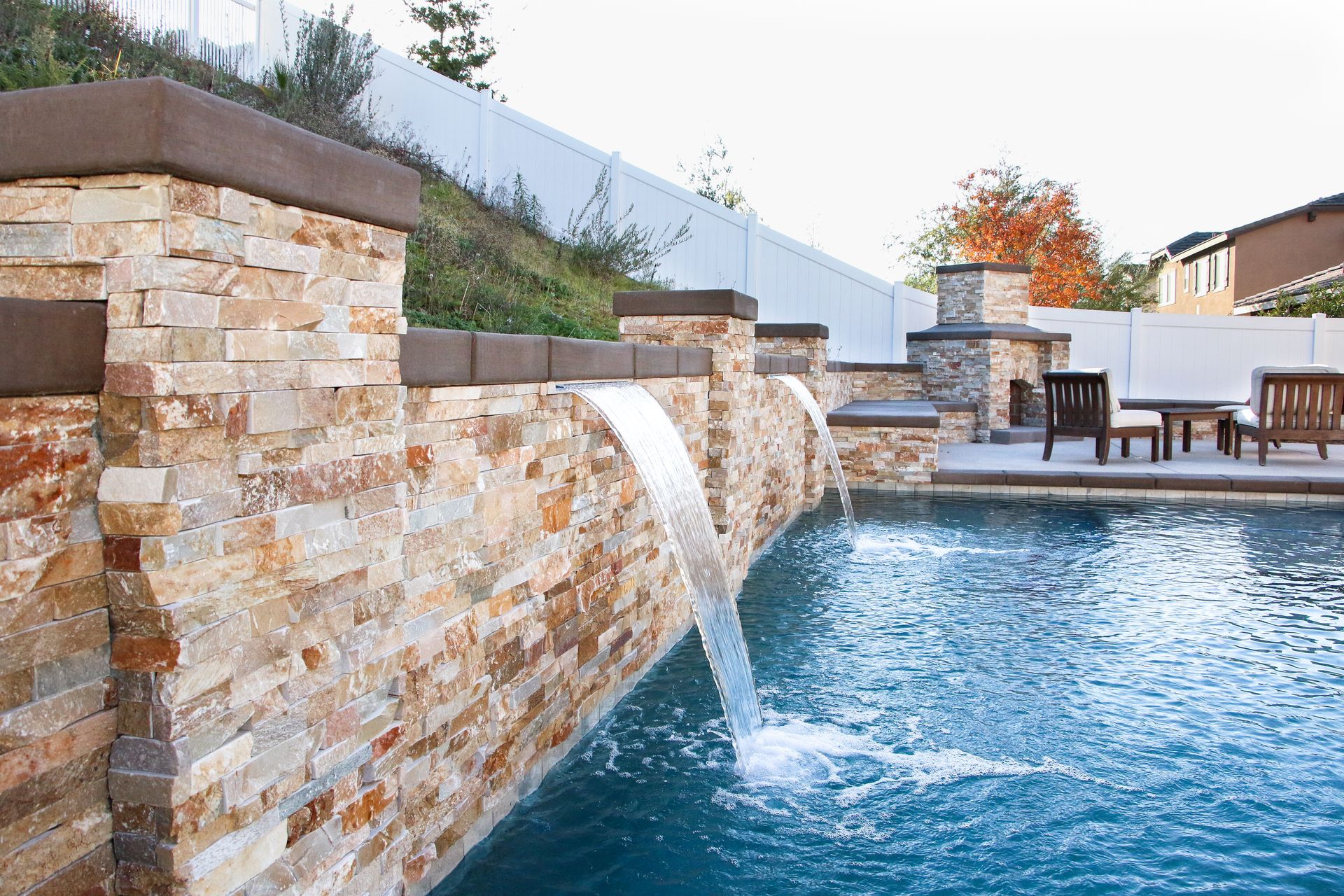 A stone retaining wall with water features pouring into a backyard swimming pool next to a patio with outdoor furniture.