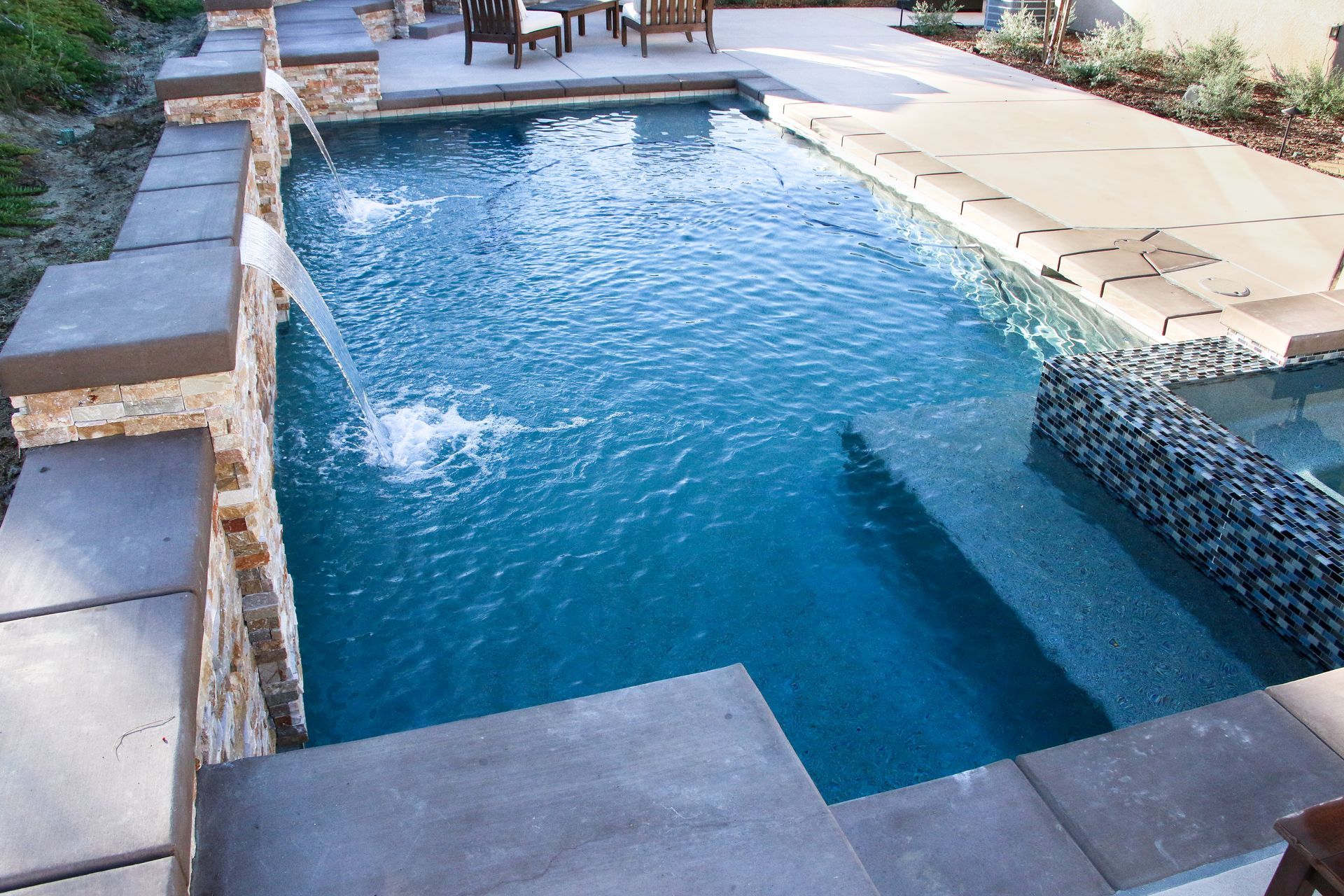 A rectangular backyard pool with a stone wall featuring cascading water features and a raised seating area.