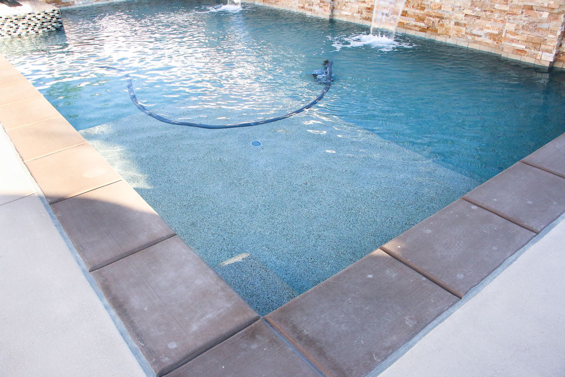 A corner of a swimming pool with dark stone pavers, clear blue water, and a pool cleaning hose visible on the step.