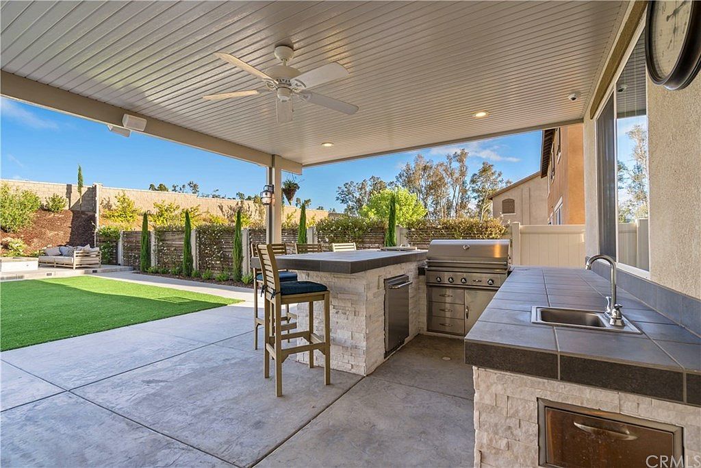 An outdoor covered patio featuring a stone kitchen island with a built-in grill, sink, and bar stool overlooking a lawn.