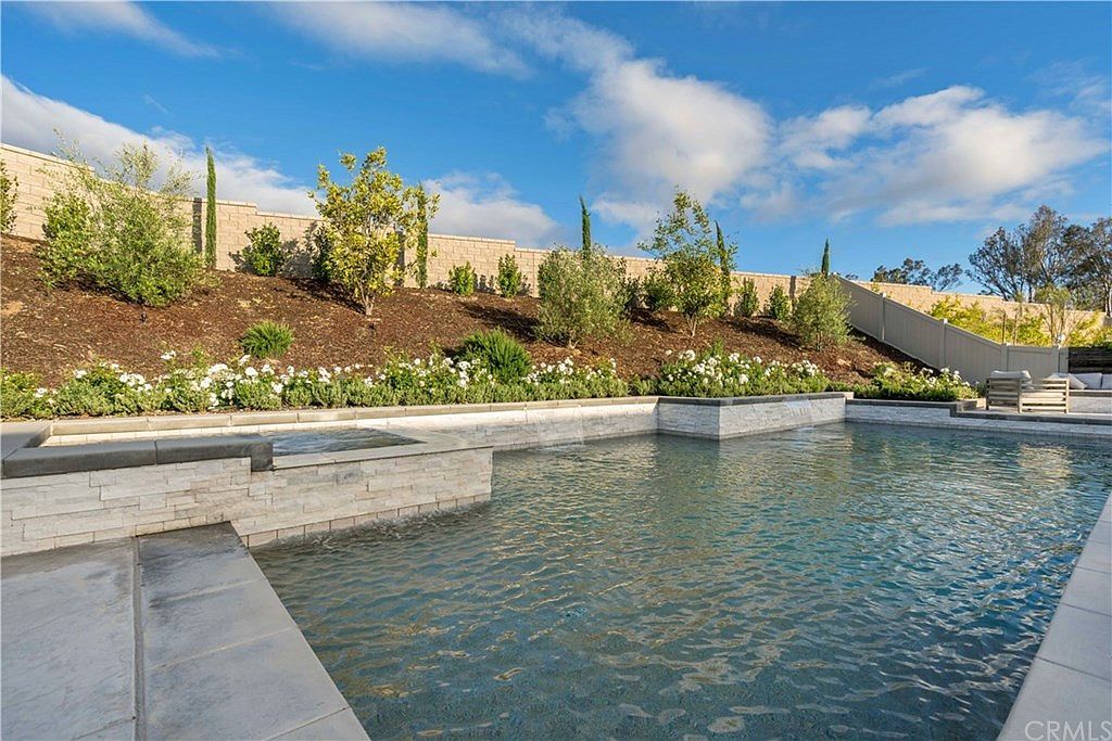 A backyard swimming pool with a stone wall feature, surrounded by landscaped terraced gardens under a blue sky.