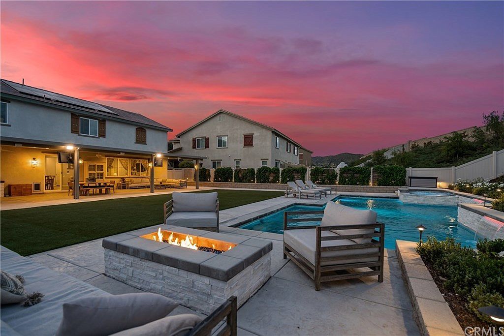 Backyard patio with a fire pit, cushioned lounge chairs, and a swimming pool at sunset with a pink and orange sky.
