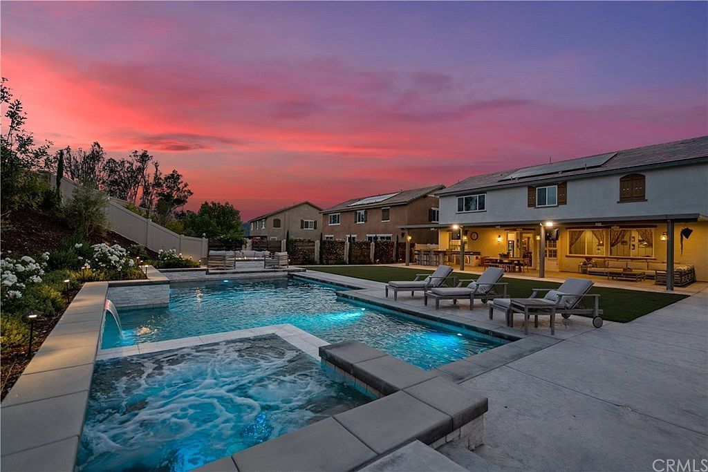 A backyard swimming pool and hot tub at twilight with a vibrant sunset sky behind a two-story suburban house.
