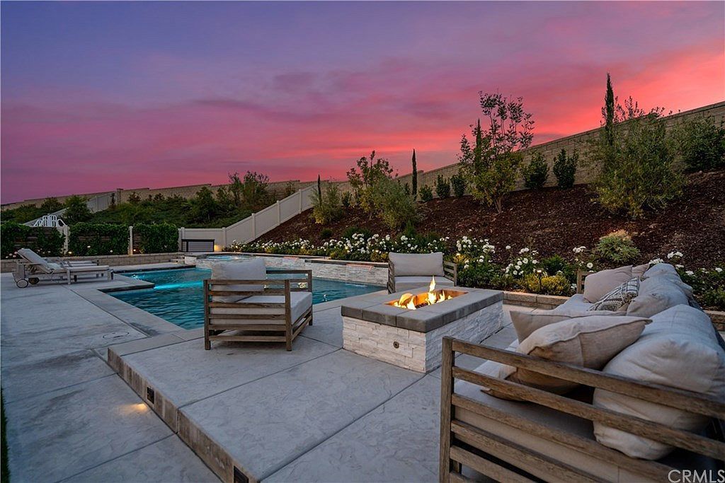A backyard patio with a fire pit, lounge chairs, and a pool at sunset with a pink and purple sky.