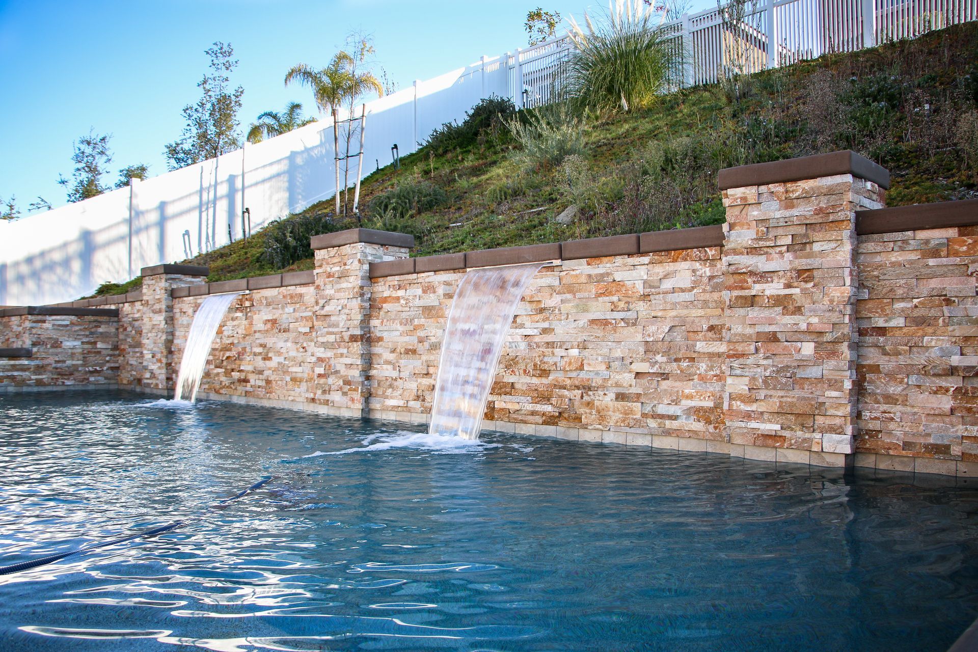 A stone retaining wall with two water features flowing into a swimming pool below a hillside.