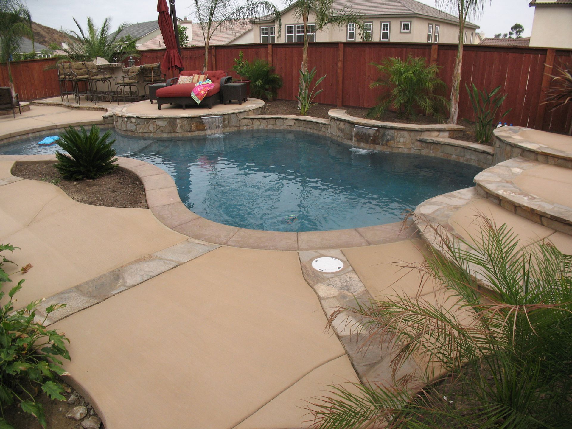 A backyard pool with a tan concrete deck, stone coping, waterfalls, and landscaping, set against a wooden privacy fence.
