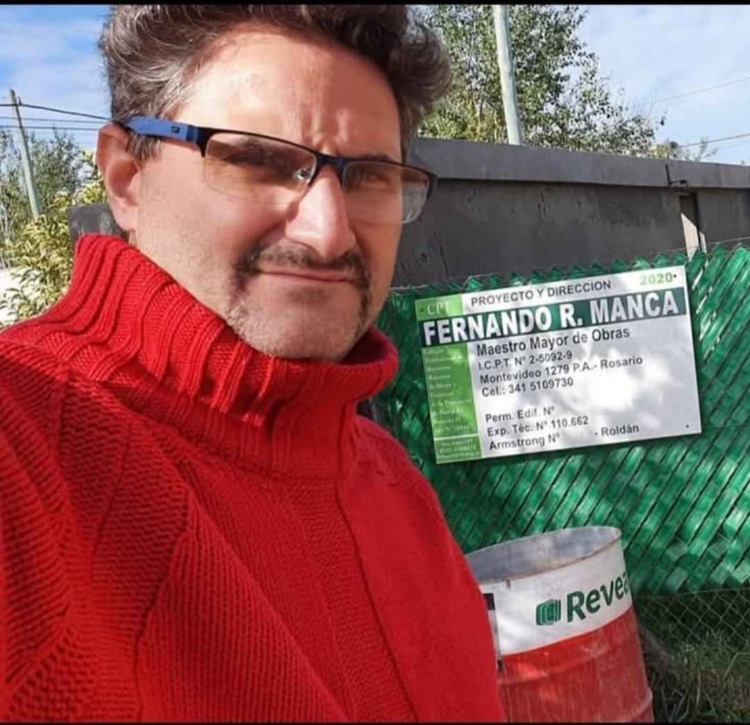 Hombre con suéter rojo, gafas, de pie cerca de un cartel con el nombre 