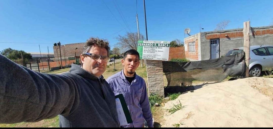 Dos hombres posan para una selfie frente a un cartel, un edificio y un automóvil en un día soleado.